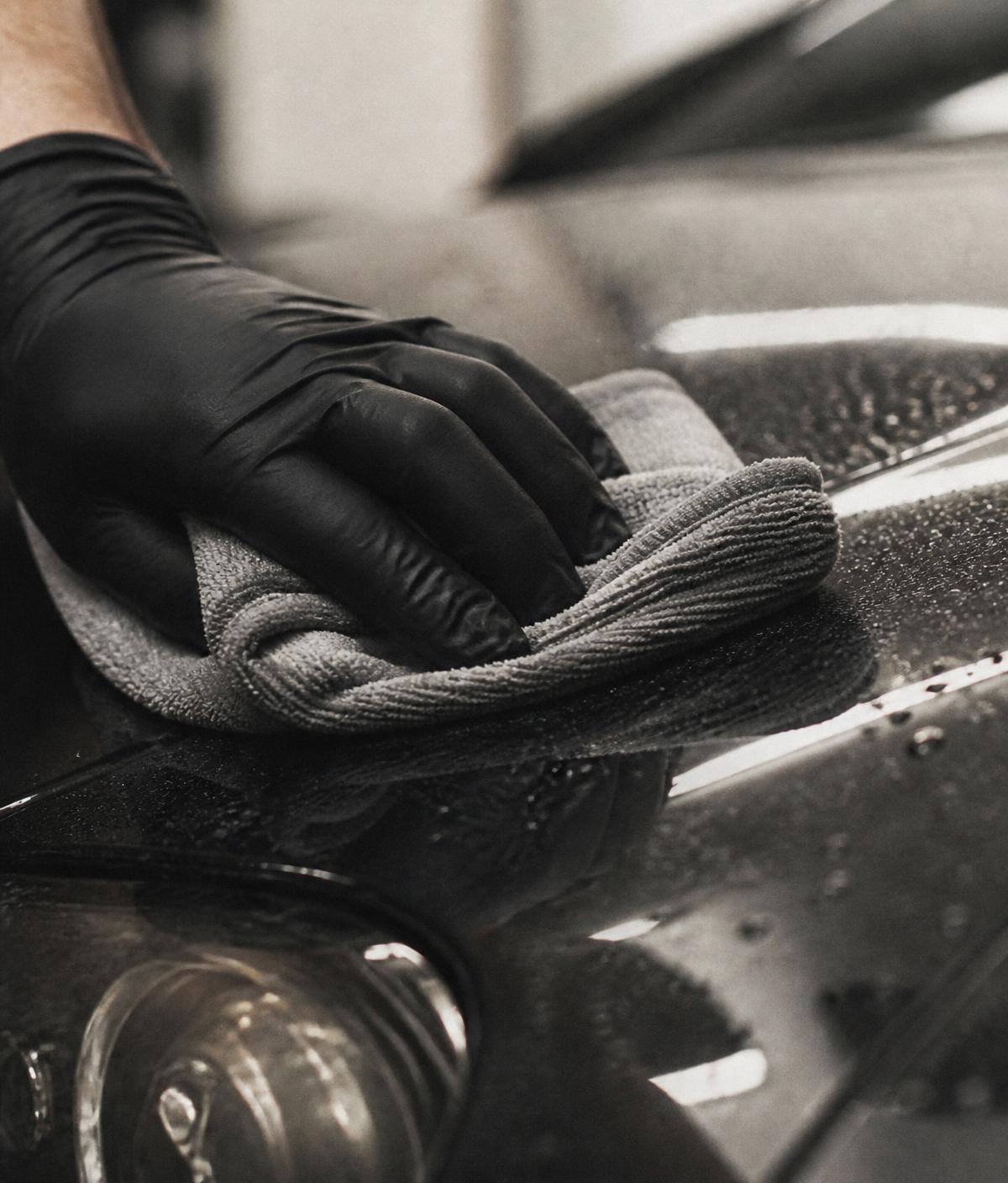 Detailer in a black nitrile glove wiping a wet charcoal car panel with microfiber
